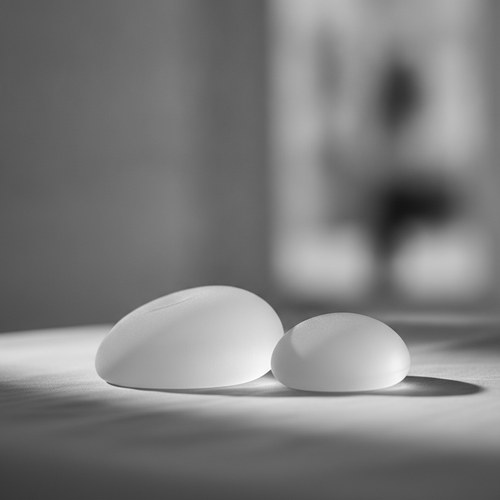 Black and white study of a woman's torso in soft light, hand resting near the collarbone. Breast implant revision commentary by Dr. Frank Agullo, MD, FACS.
