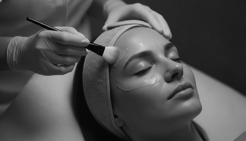 Black and white close-up of a patient reclining with eyes closed while a practitioner in pink gloves applies peel solution to her cheek with a fan brush. Chemical peel editorial by Dr. Frank Agullo, MD, FACS.