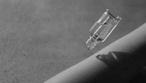 Black and white editorial close-up of a dropper bottle of serum against bare skin, illustrating hyaluronic acid serum commentary by Frank Agullo, MD, FACS.