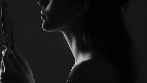 Moody greyscale photograph of a woman in profile examining her jawline and under-chin in a handheld mirror. Kybella fat-dissolving injection review by Frank Agullo, MD, FACS, Dr. WorldWide.
