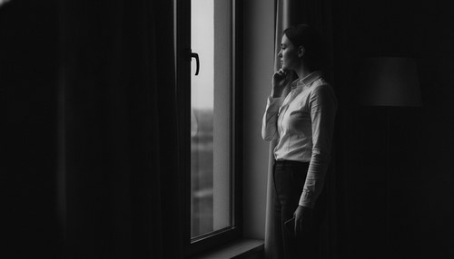Black and white photograph of a woman in profile at a tall dark window at dusk, one hand resting at her jaw, face turned to the glass. Illustrative image, not a patient. Commentary by Frank Agullo, MD, FACS.