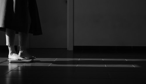 Black and white photograph of a bathroom scale on a dark tiled floor beside a doorway, a figure's feet and the hem of a robe at the edge of the frame, hard side light, no face. Illustrative image, not a patient. Commentary by Frank Agullo, MD, FACS.
