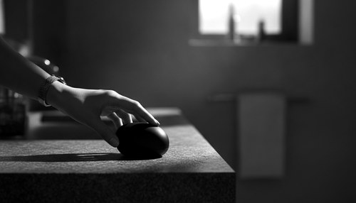 Black and white photograph of a hand resting a small unbranded handheld skin device on a dark stone bathroom counter, hard side light, no face in frame. Illustrative image, not a patient. Commentary by Frank Agullo, MD, FACS.