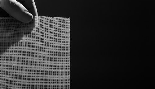 Black and white close-up photograph of a sheet of fine surgical mesh held up against hard side light so the weave shows, dark background, gloved fingertips at the edge of the frame, no face. Illustrative image. Commentary by Frank Agullo, MD, FACS.