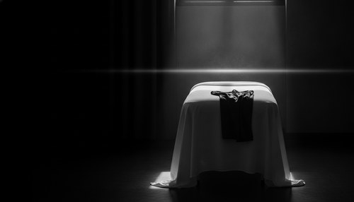 Black and white photograph of a massage table in a dim treatment room, white linen, a folded compression garment at its foot, a single shaft of window light, no person. Illustrative image, not a patient. Commentary by Frank Agullo, MD, FACS.