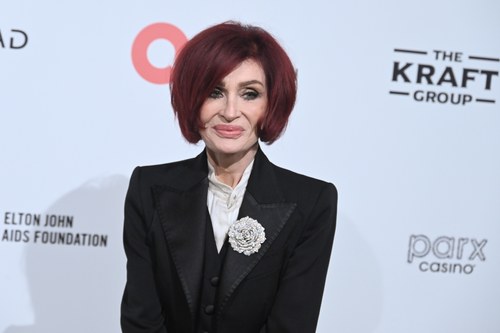 Sharon Osbourne on the red carpet in March 2026, deep red bob, black tuxedo jacket with a crystal flower brooch, facing the camera. Commentary by Frank Agullo, MD, FACS.